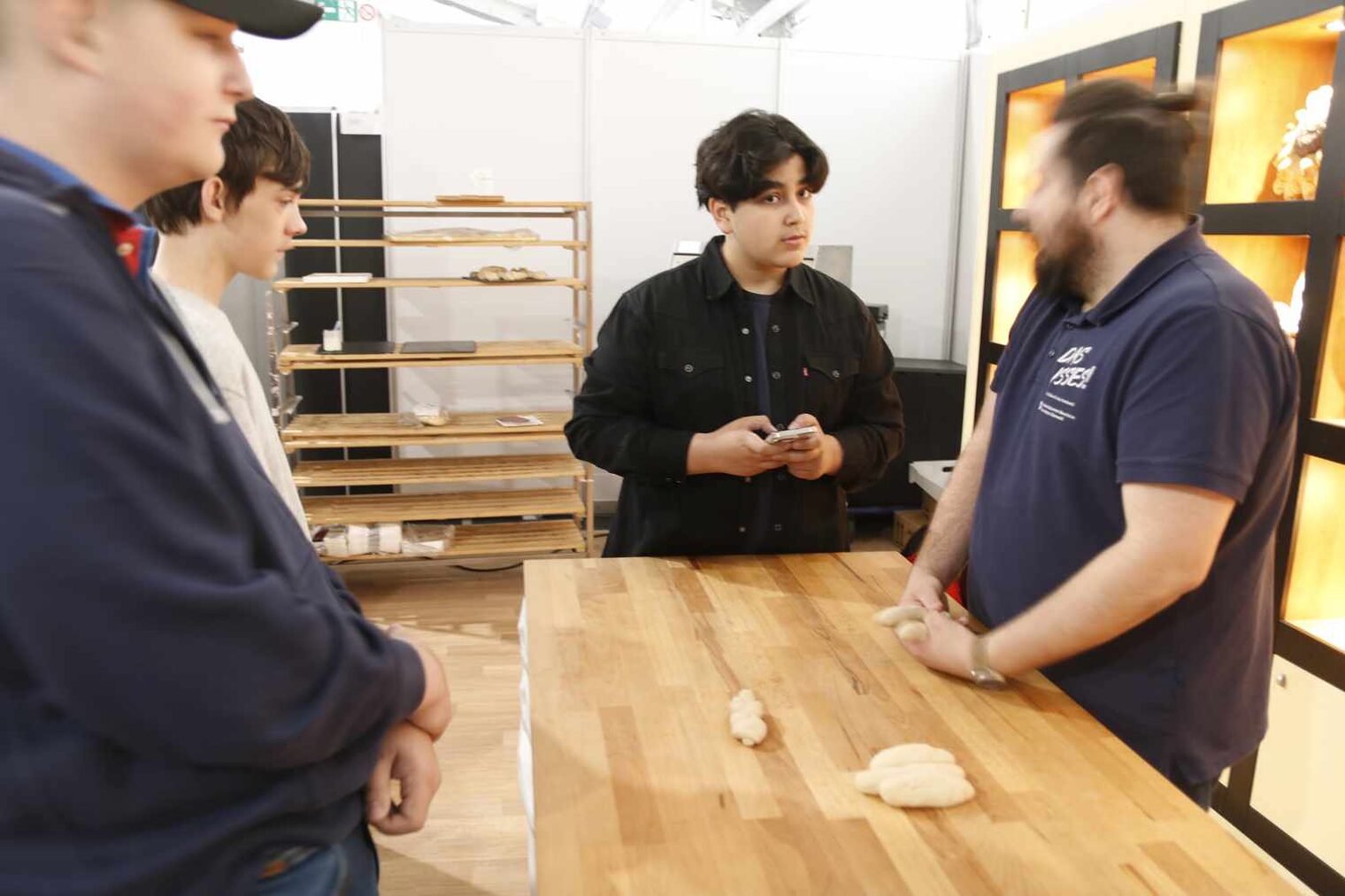 Drei Jungs stehen an einem bemehlten Tisch mit Teigrollen und sind in ein Gespräch mit dem Bäcker vertieft, ein Junge mit Smartphone.
