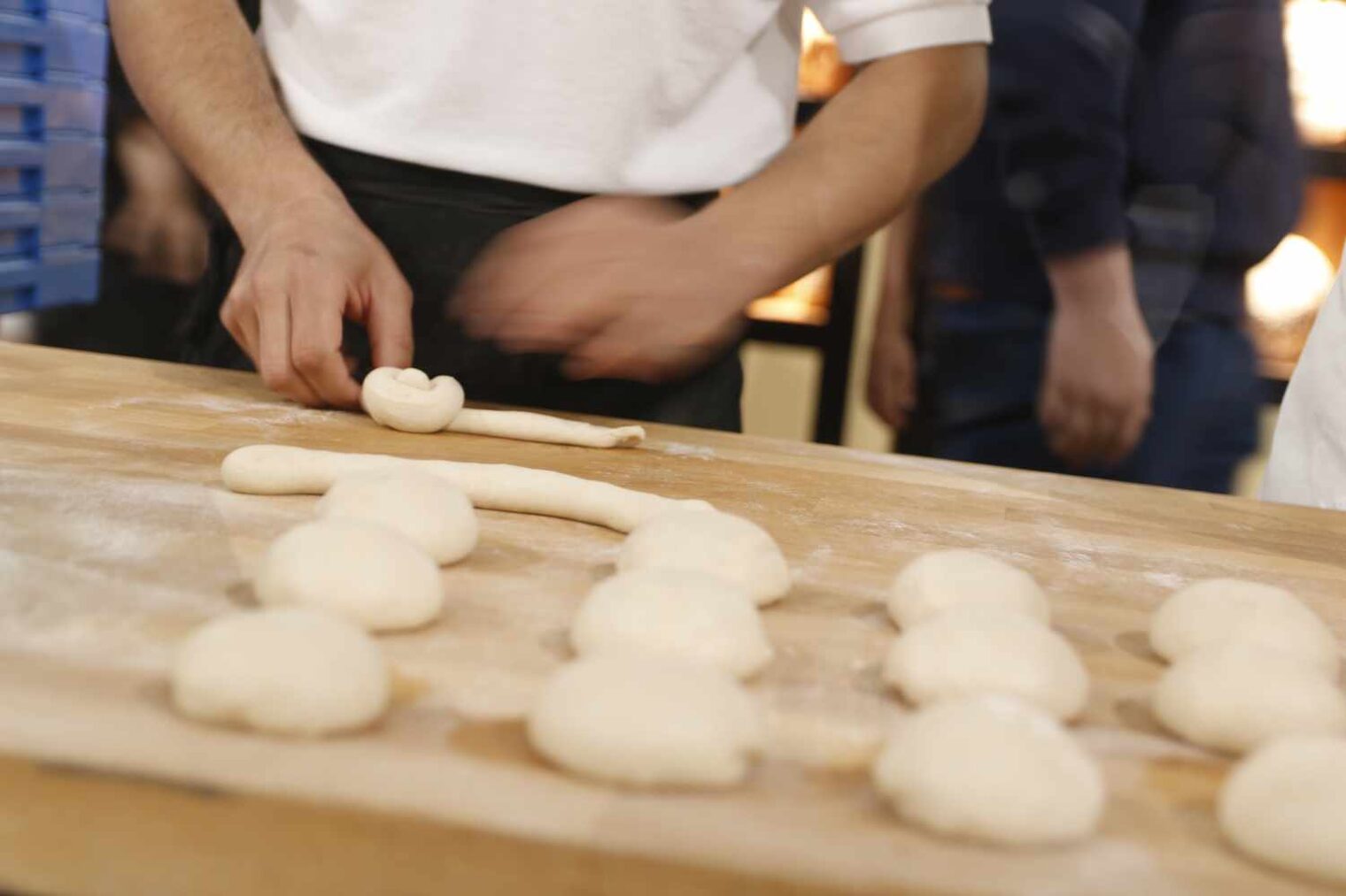 Nahaufnahme eines Holztisches mit Teig, der in Brötchenformen geformt ist, im Hintergrund leicht verschwommen die Hände eines Bäckers
