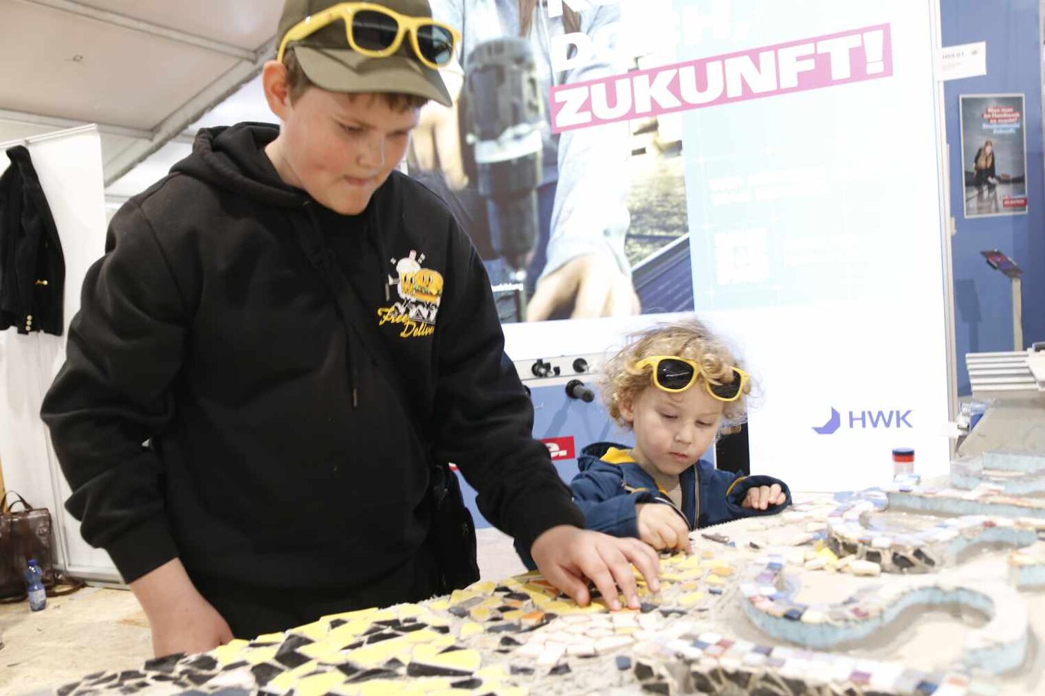 13-jähriger Junge verlegt auf einer liegenden Tafel bunte Fliesenmosaike, daneben ein kleines Kind mit blonden Locken und Sonnenbrille