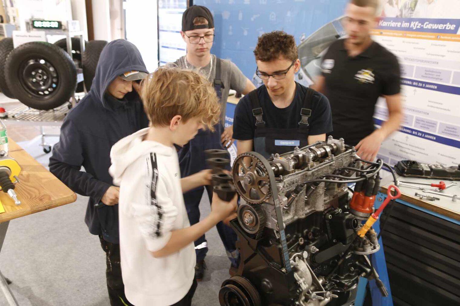 Vier Schüler stehen an einem Motor und begutachten, wie dieser funktioniert; leicht verschwommen rechts im Hintergrund ein Kfz-Mechaniker