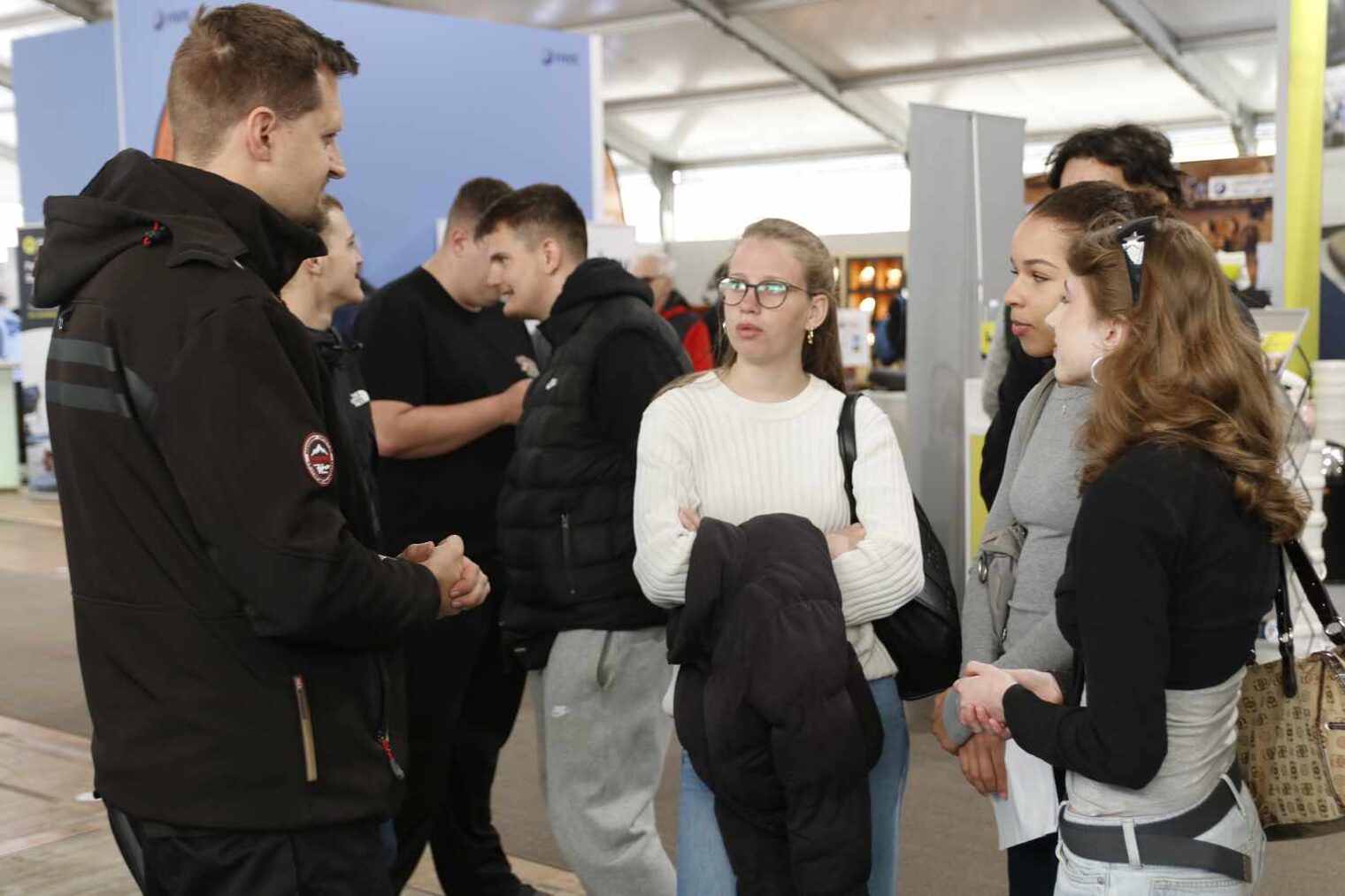 Mann am linken Bildrand im Gespräch mit drei Schülerinnen, die aufmerksam zuhören; im Hintergrund Jungs, die Blödsinn machen