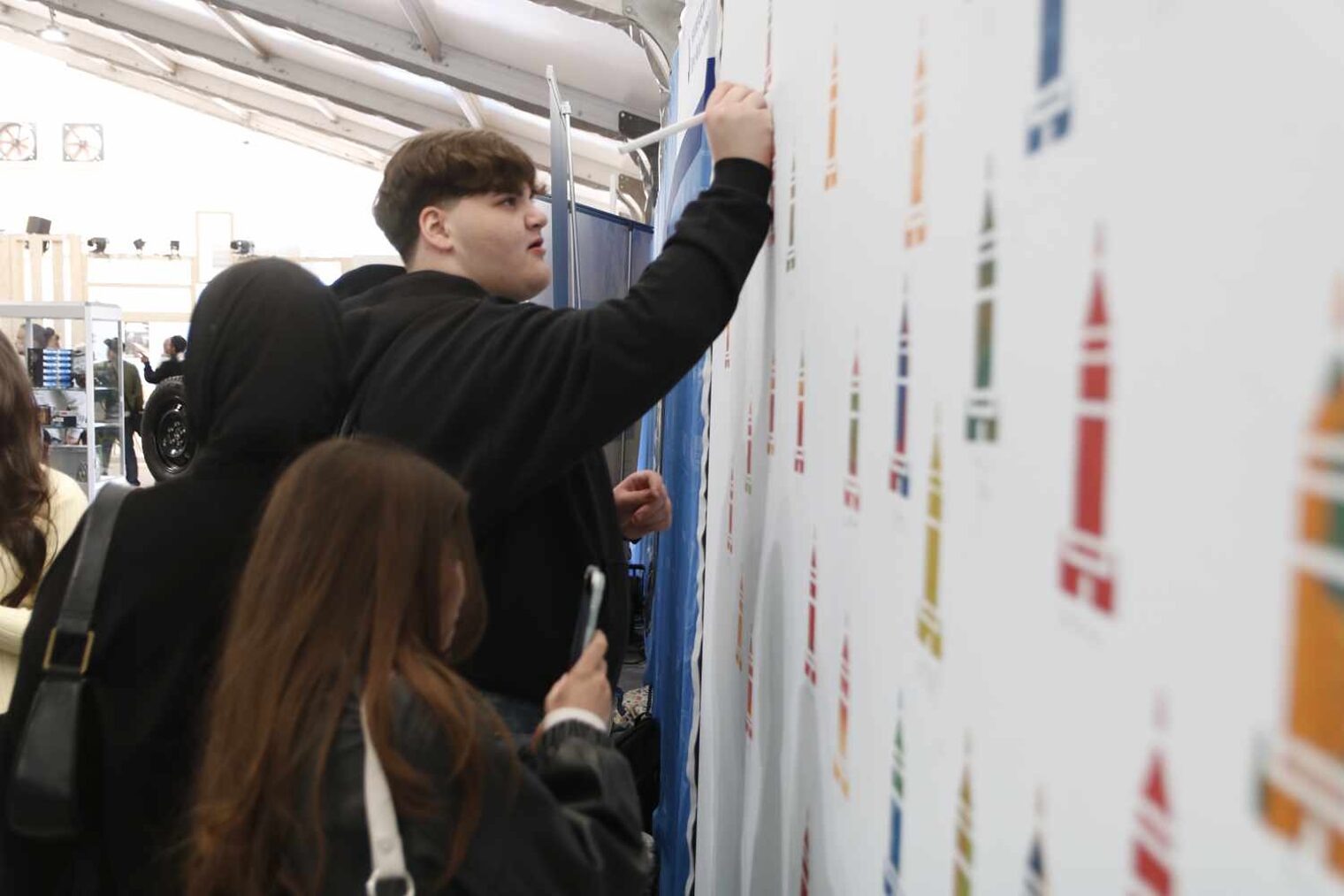 Schüler vor einer weißen Wand mit lauter bunten Turm-Motiven malt mit einem Pinsel selbst etwas auf, davor schauen zwei Mädchen zu