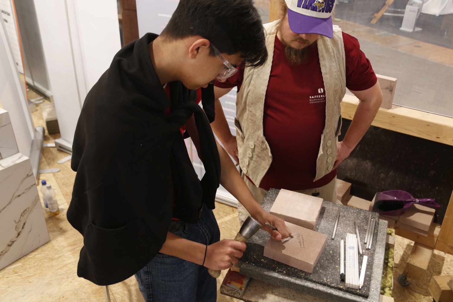 Schüler bearbeitet auf kleinem Tisch eine Steinplatte mit dem Meißel, rechts von ihm ein Steinmetz, der die Handgriffe erklärt