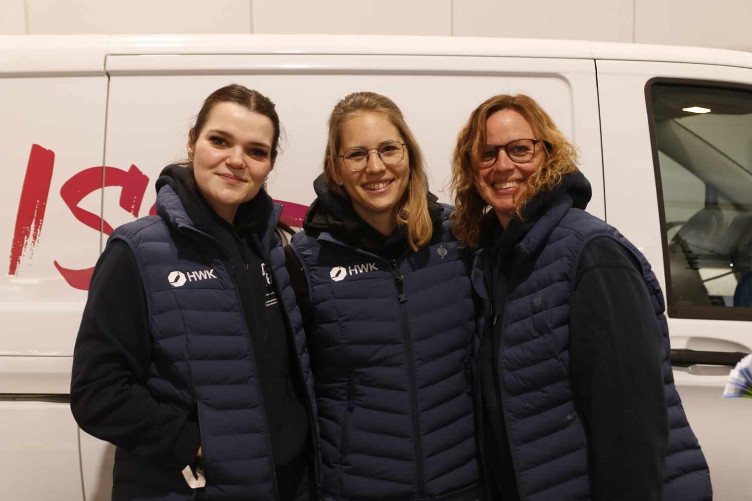 Drei junge Frauen, gleich gekleidet mit blauen Hoodies und Steppwesten, vor der Silhouette eines weißen Transporters mit Schriftzug.