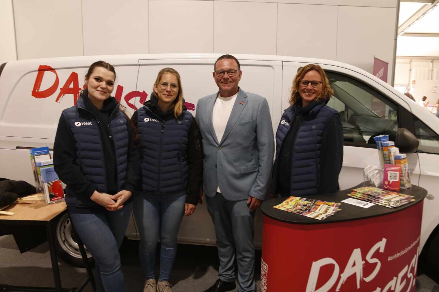 Drei junge Frauen - zwei links, eine rechts - neben einem Mann im hellblauen Stoffanzug vor einem weißen Transporter; rechts vorne ein Desk