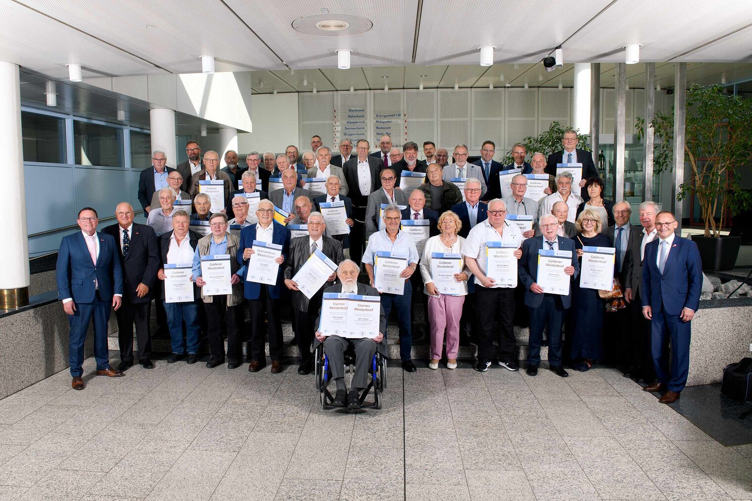 Große Gruppe von Senioren in einer Festsaal-Aula, jeweils mit Ehrenurkunden in den Händen, ganz vorne in der Mitte ein Herr im Rollstuhl