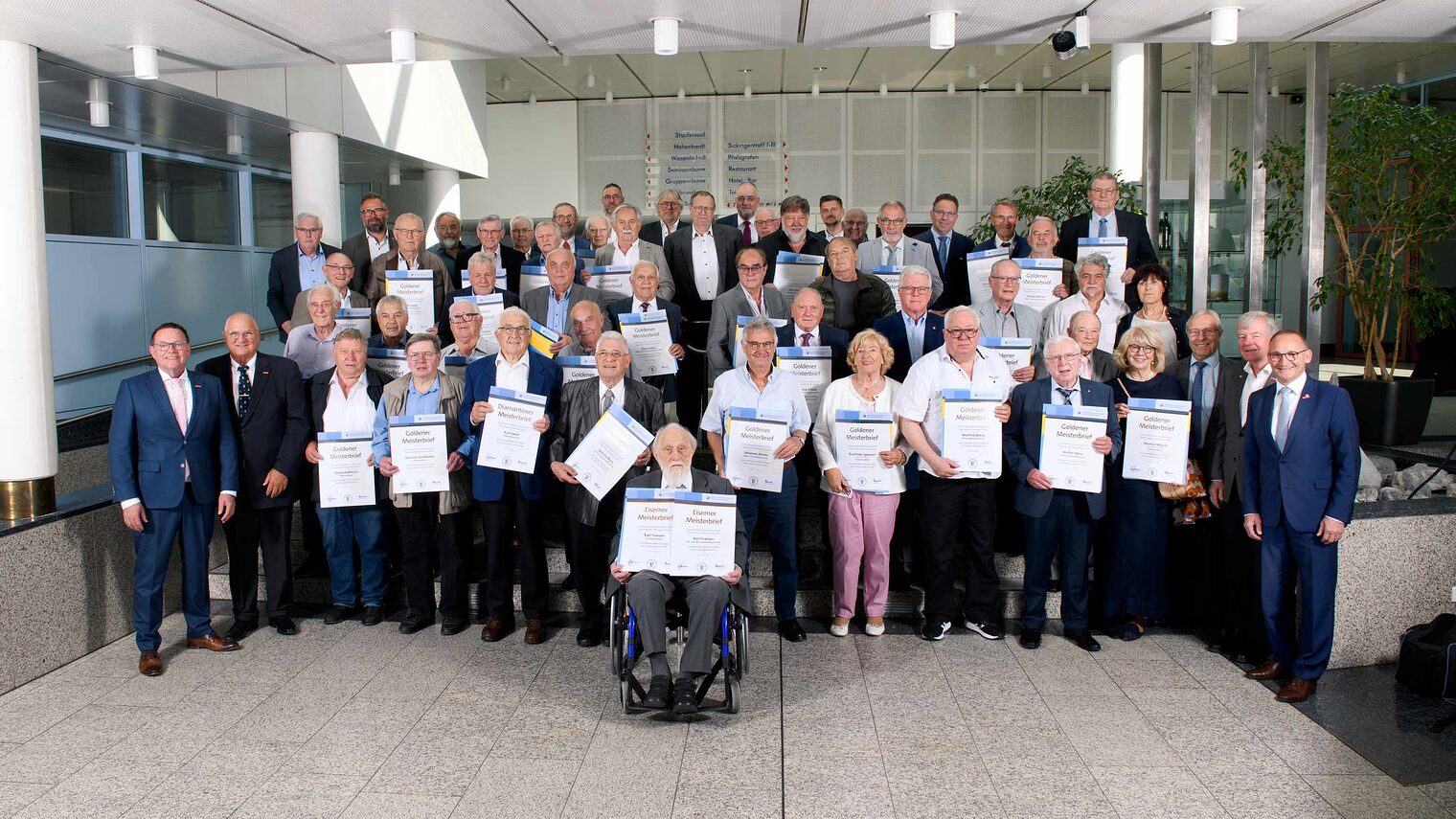 Große Gruppe von Senioren in einer Festsaal-Aula, jeweils mit Ehrenurkunden in den Händen, ganz vorne in der Mitte ein Herr im Rollstuhl