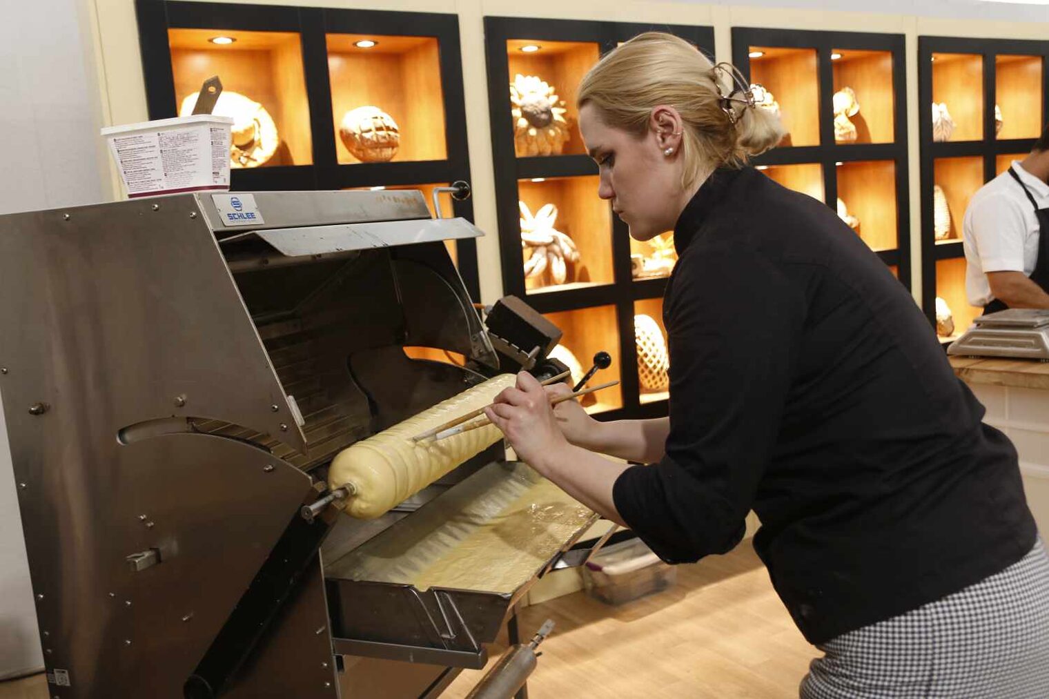 Blonde Frau in traditioneller Bäckerkluft vor dem Spezialofen, in dem sie einen Baumkuchen mit Stäben zur typischen Form bearbeitet