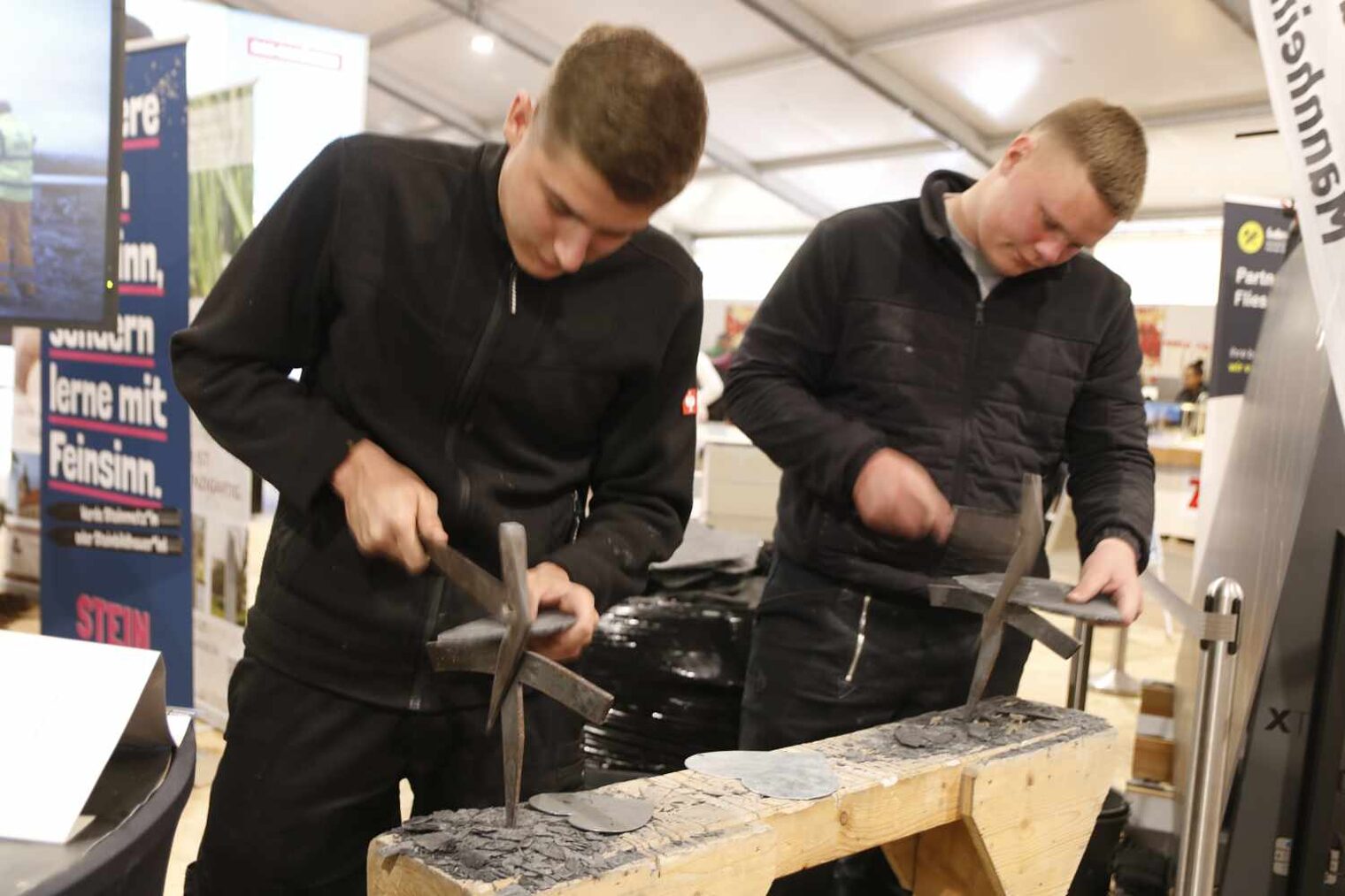 Zwei Handwerker in schwarzer Dachdecker-Kluft an einem schmalen Holzbalken mit Amboss arbeiten mit Werkzeugen an Schiefertafeln
