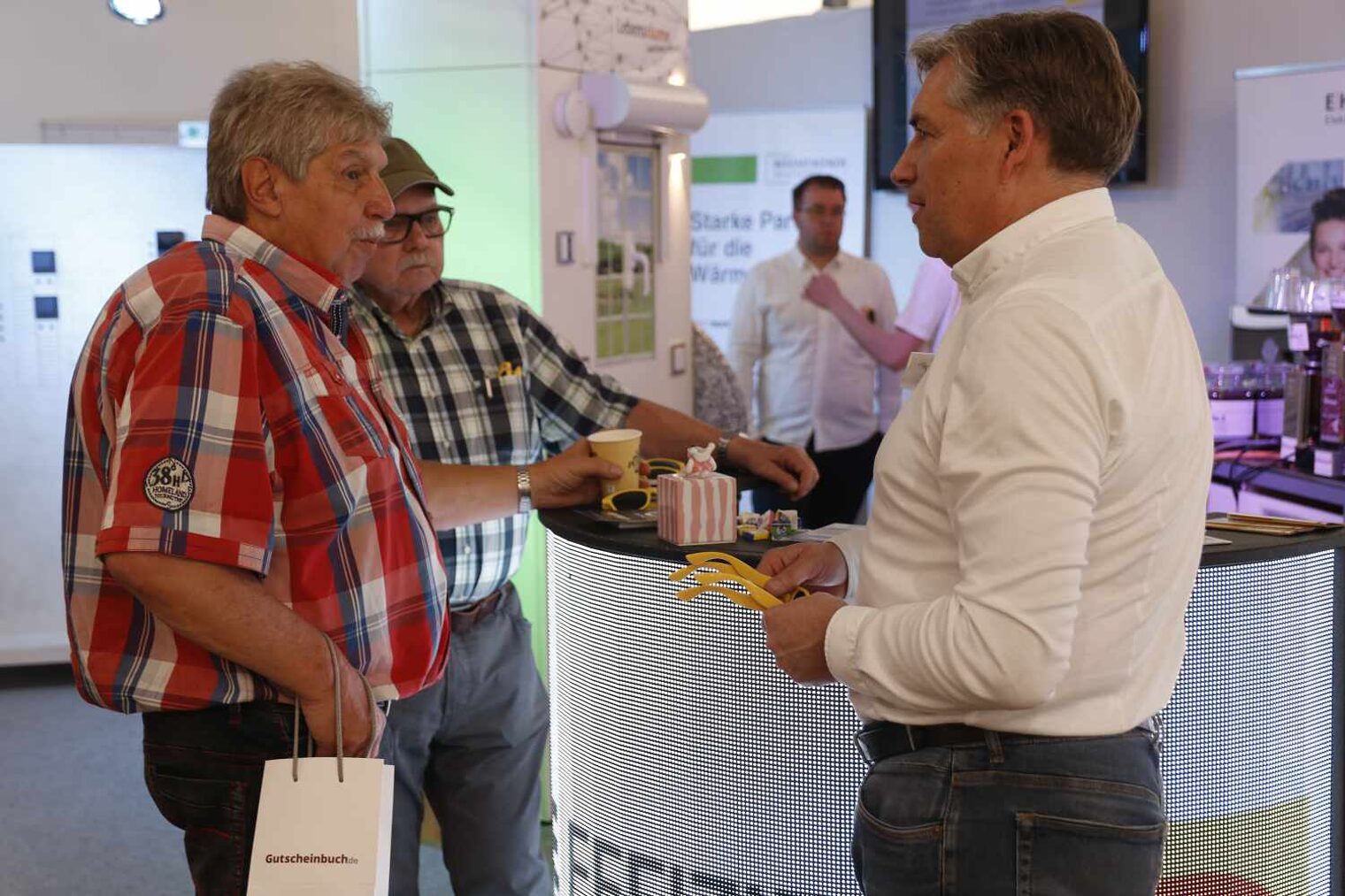Drei Männer an einem Messestand, vertieft in ein Gespräch; die beiden ältern Männer links in karierten Hemden, der mann recht in Weiß