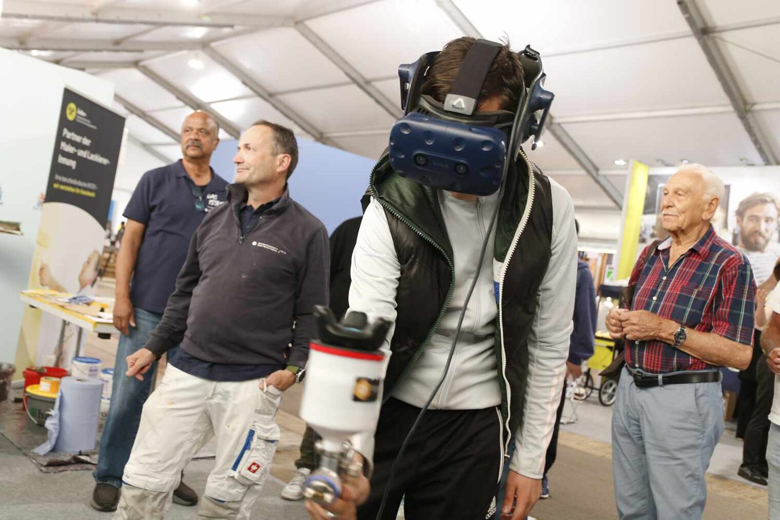 Junger Mann, dessen Kopf eine VR-Brille bedeckt, streckt eine Spritzpistole entgegen; im Hintergrund weitere Männer, die zusehen