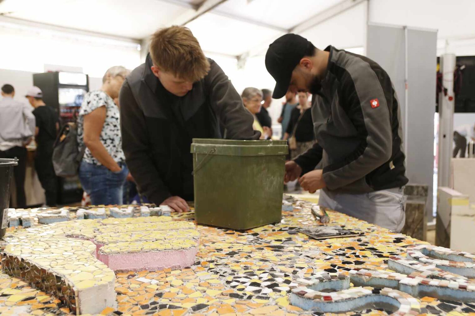 Zwei Jungs arbeiten an einer großen Platte mit Fliesenmosaiken, vor ihnen ein großer Eimer, beide sind vertief in ihre Arbeit