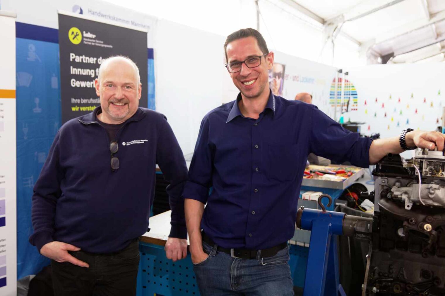 Zwei Männer mit dunkelblauen Oberteilen an einem Messestand, der rechte legt die Hand auf eine Ablage und trägt Brille