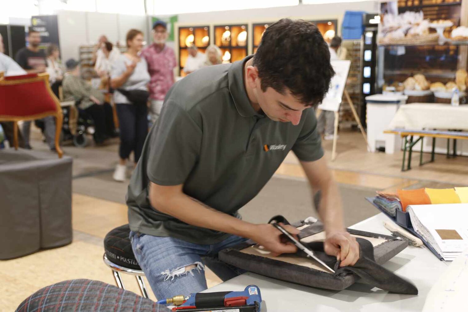 Sattler arbeitet an einem Messestand an der Polsterung eines Stuhlsitzes über den er gebeugt sitzt und ein Werkzeug in der Hand führt