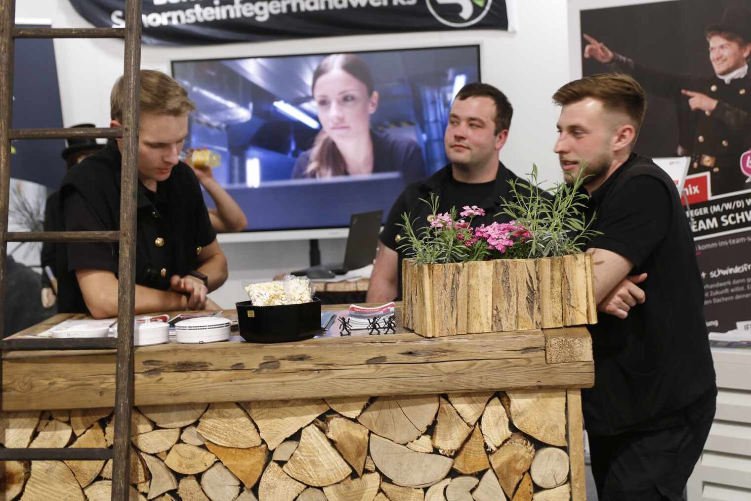 Drei Männer in schwarzen Schornsteinfeger-Outfits hinter einer Theke im Look eines Holzstapels, seitlich lehnt eine Holzleiter