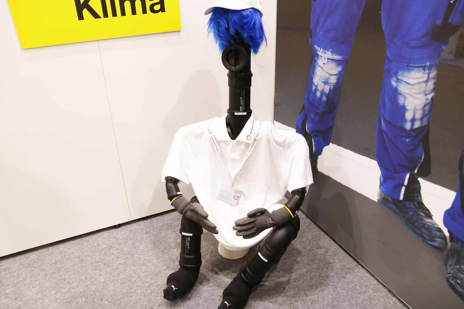Sitzendes Männchen aus schwarzen Plastikrohren gebastelt mit einer blauen Haar-Perücke und weißem Cap und T-Shirt in der Ecke eines Messestands