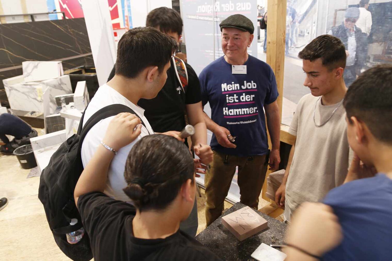 Gruppe aus fünf jugendlichen Männern und einem älteren Mann mit Baskenmütze steht kreisförmig an einem Messestand und unterhält sich