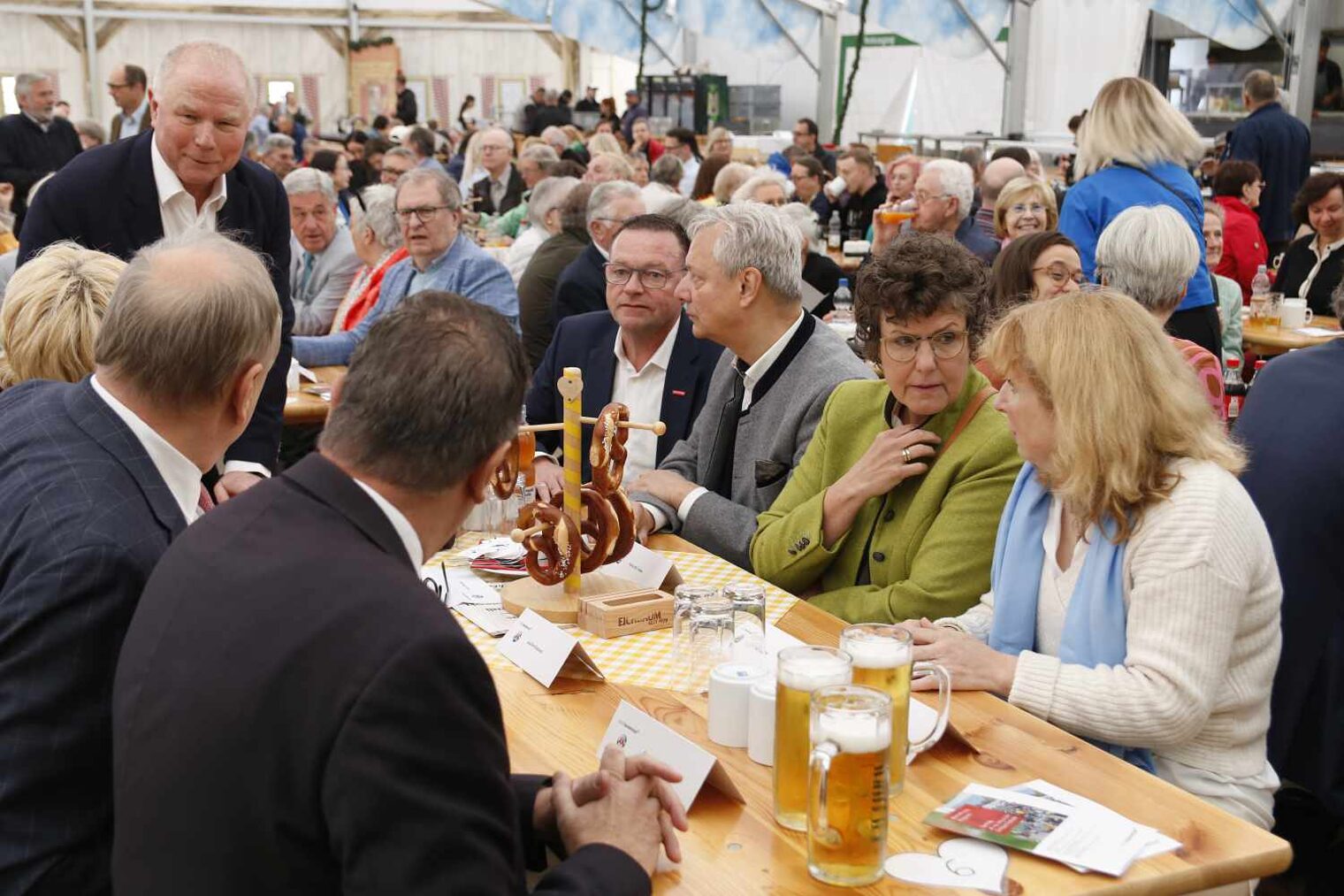 Szene im Bierzelt, vorne sitzen an einem Holztische Männer und Frauen im Gespräch vertieft, auf dem Tisch stehen Bierkrüge und Brezelhalter