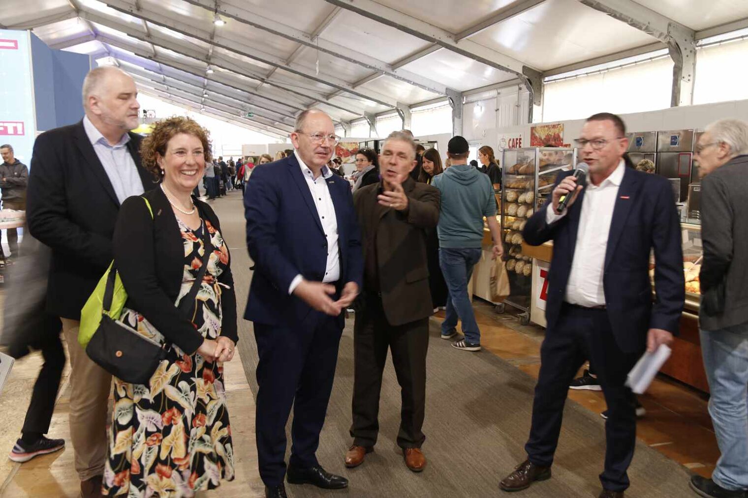 Personen laufen durch Messezelt, vorne eine lachende Frau im gemusterten Kleid, in der Mitte zwei Männer, wovon einer zeigt, rechts ein Mann mit Mikrophon