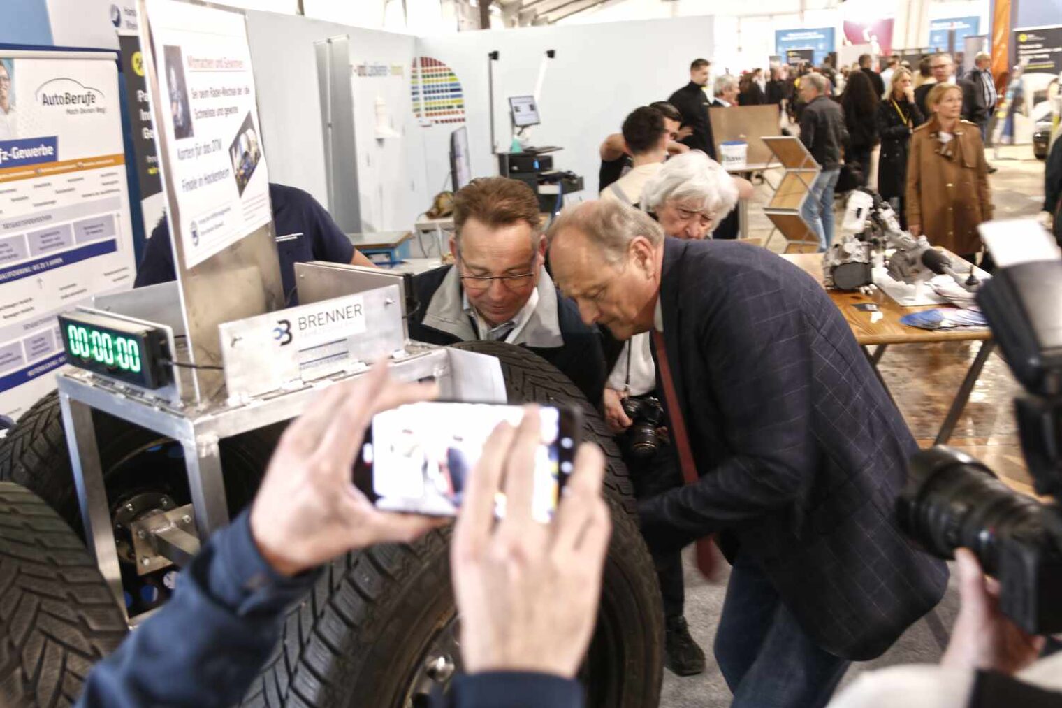 Übersicht über einen Messestand des Kfz-Handwerks mit vielen Besuchern im Hintergrund, zwei Männern an einem Reifen und hochgehaltenem Smartphone vorne