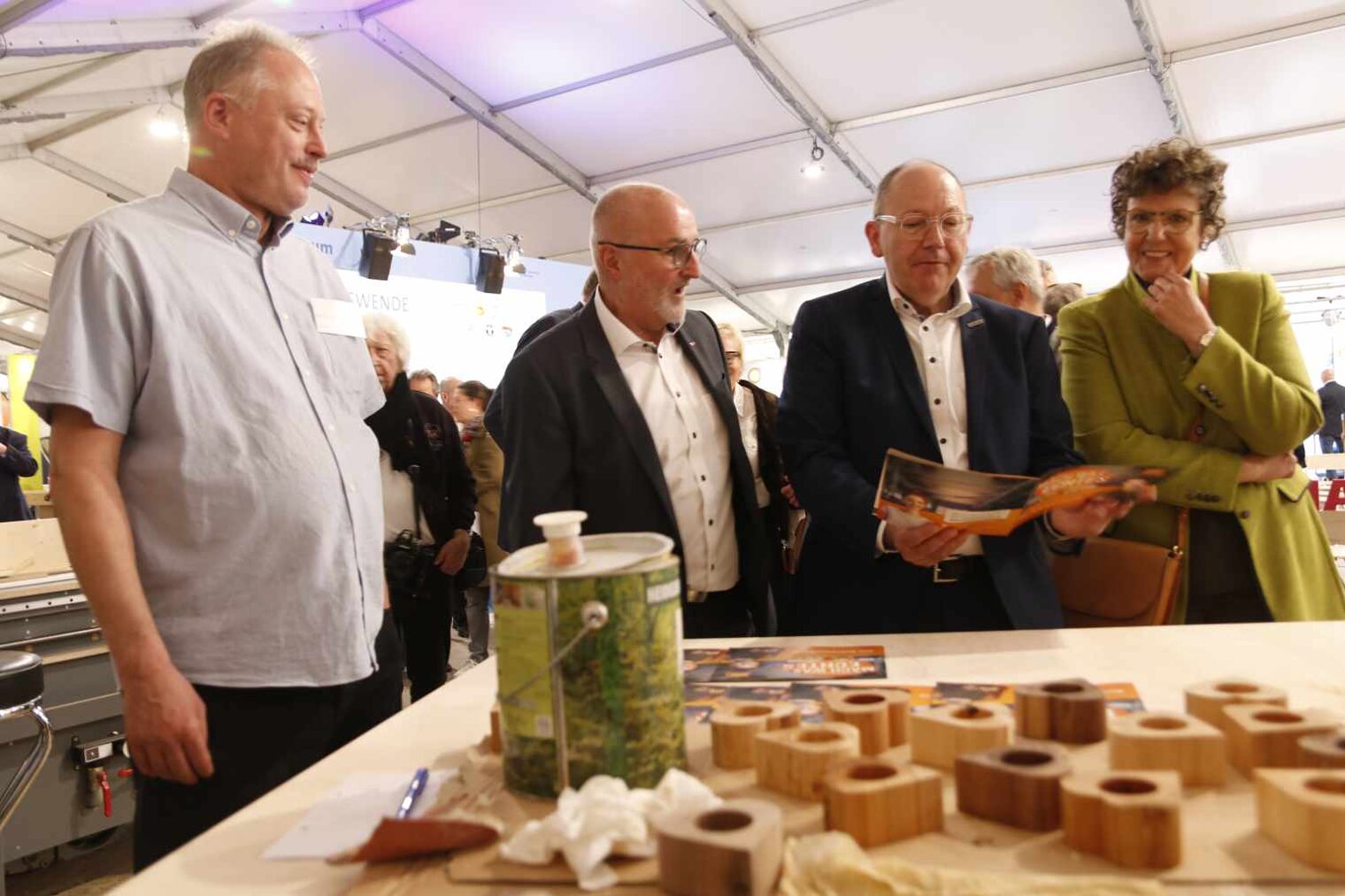 Drei Männer und eine Frau stehen an einem Tisch in einer Messehalle und betrachten die Holzarbeiten, die im Vordergrund sichtbar darauf liegen