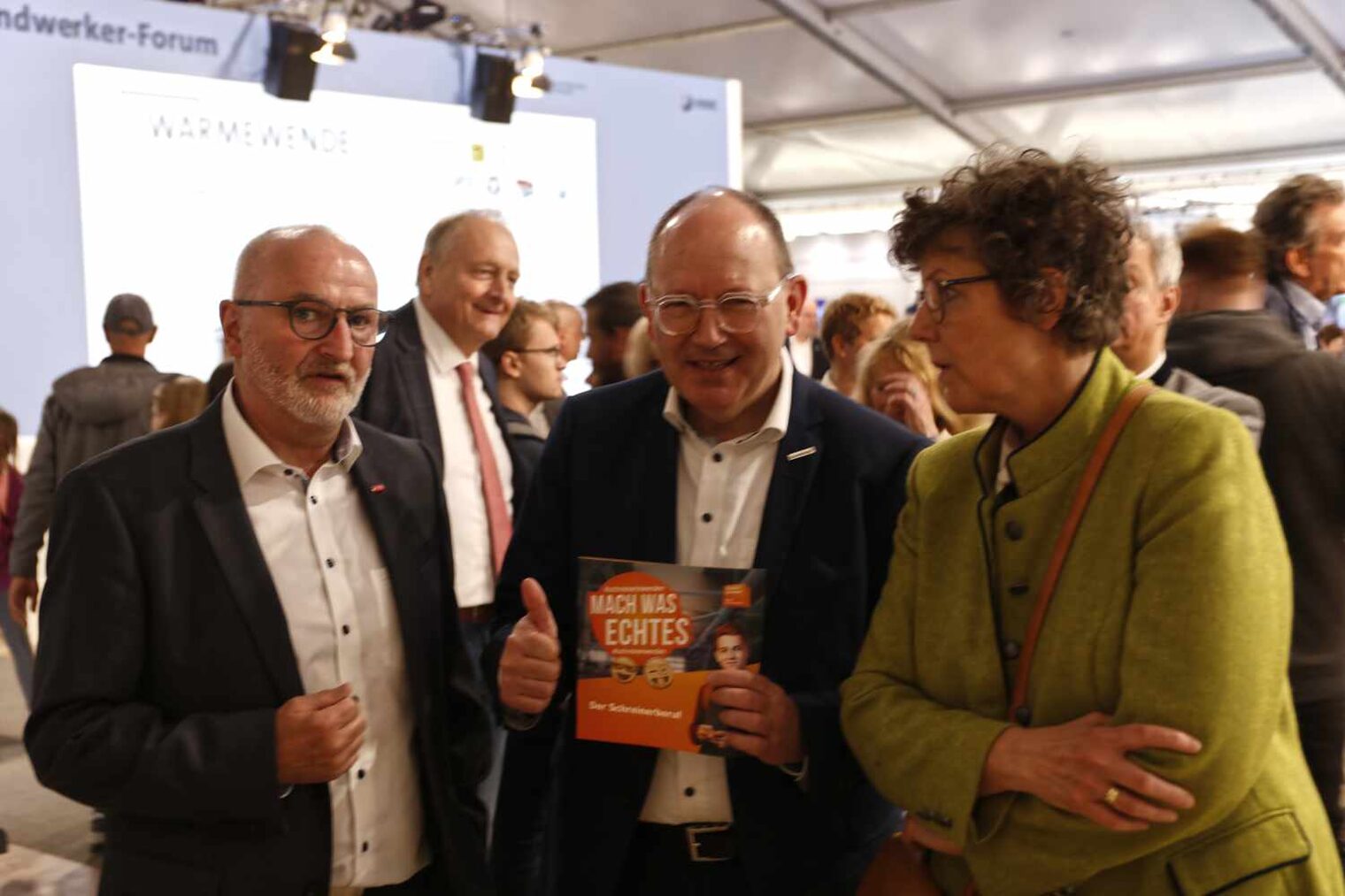 Blick auf Messegeschehen mit vielen Personen, im Vordergrund zwei Männer und eine Frau, der mittlere Mann macht das Daumen-hoch-Zeichen