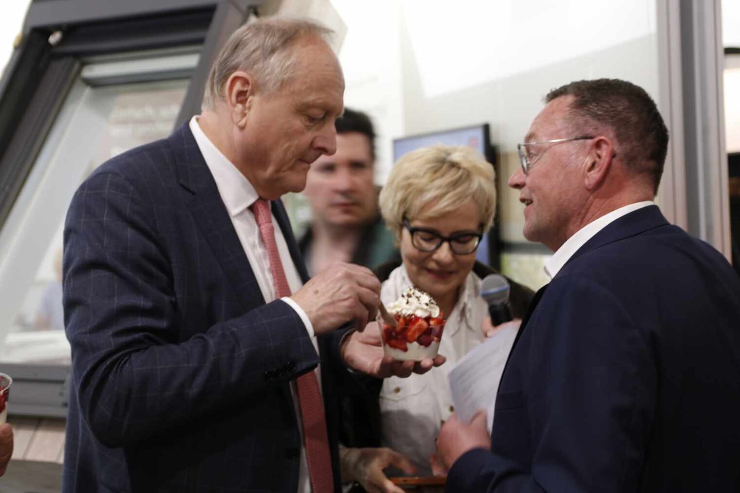 Zwei Männer im Vordergrund, der Mann rechts mit Brille redet auf den anderen ein, der einen Erdbeerbecher ist, im Hintergrund eine blonde Frau