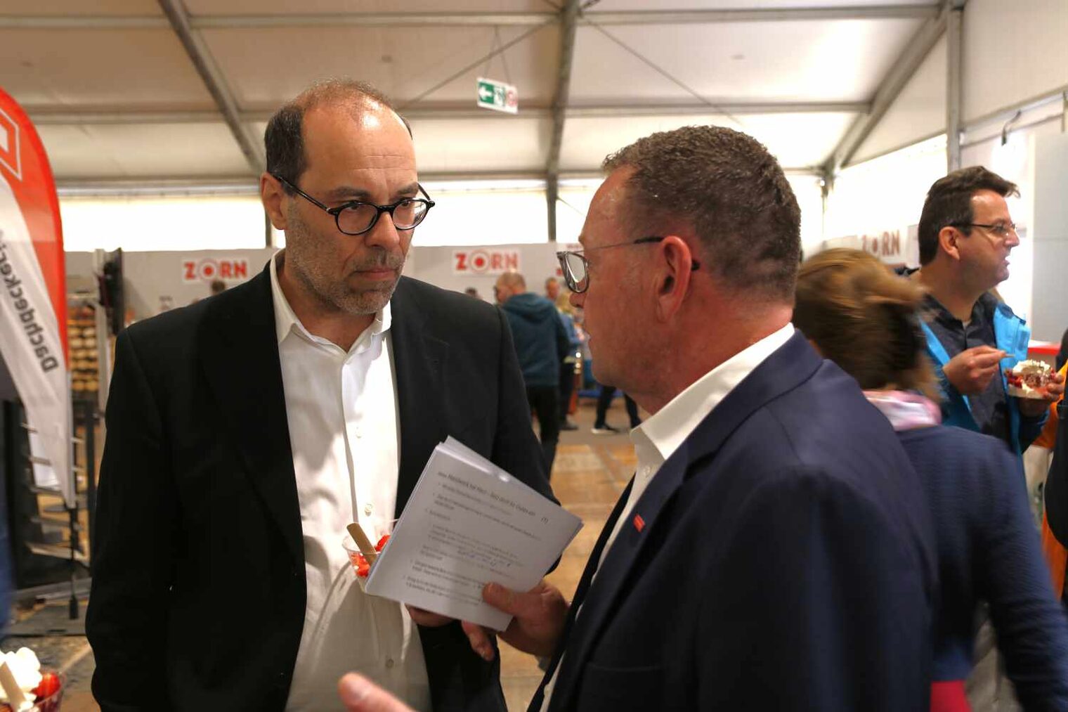 Zwei Männer in einer Messehalle im Gespräch, beide tragen Brillen, der rechts hält ein Manuskript in der Hand, im Hintergrund laufen andere