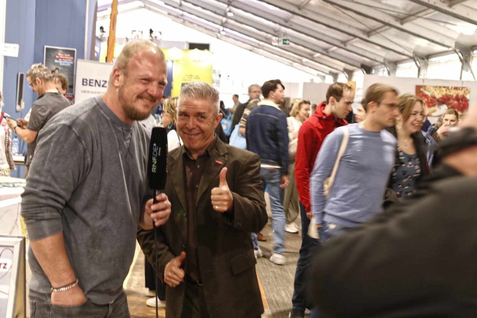 lebendige Situation in einer Messehalle mit vielen Besuchern; vorne zwei Personen, der Mann links mit Radiomikrophon in der Hand, der rechts mit Daumen hoch in die Kamera