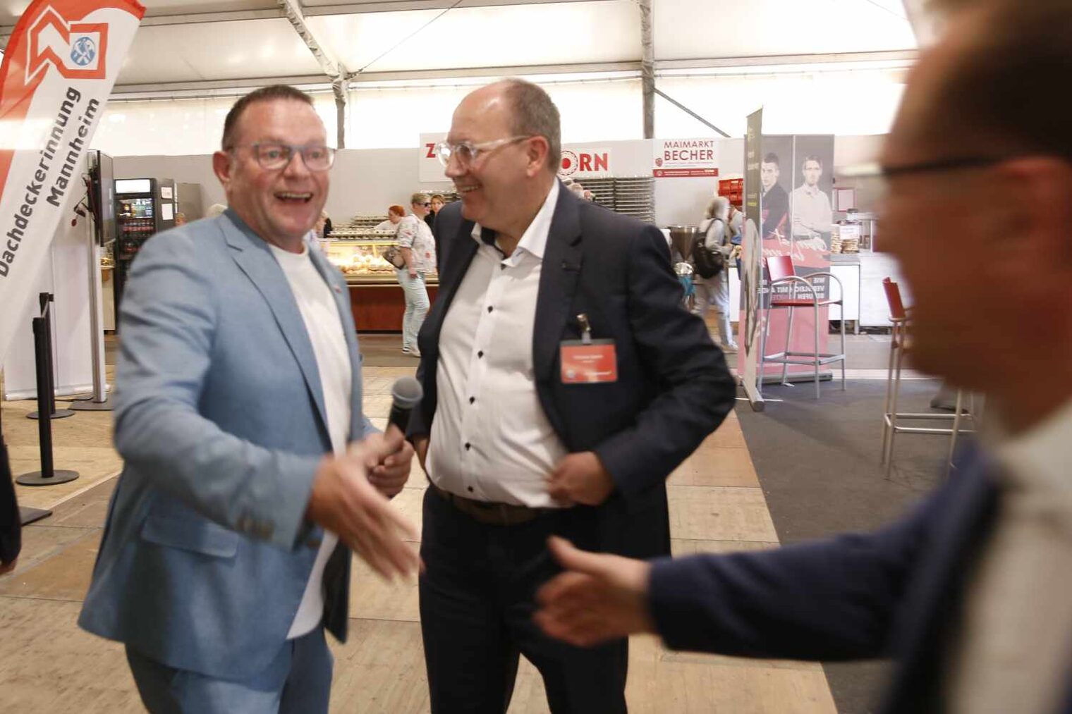 links ein Mann im hellblauen Anzug, der lachend einem weiteren Mann, der gerade ins Bild läuft, die Hand entgegenstreckt, dazwischen ein weiterer Mann
