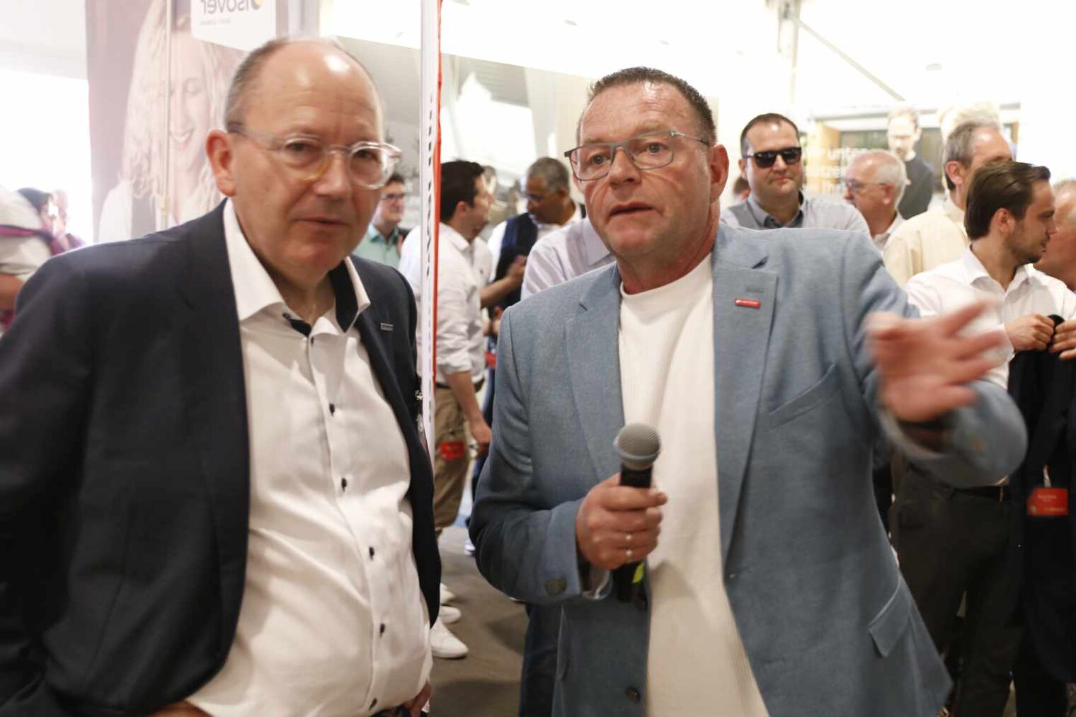 bewegte Szene in einer Messehalle, vorne zwei Männer, der links mit Brille hört zu, der rechts hat ein Mikro in der Hand und gestikuliert mit der anderen