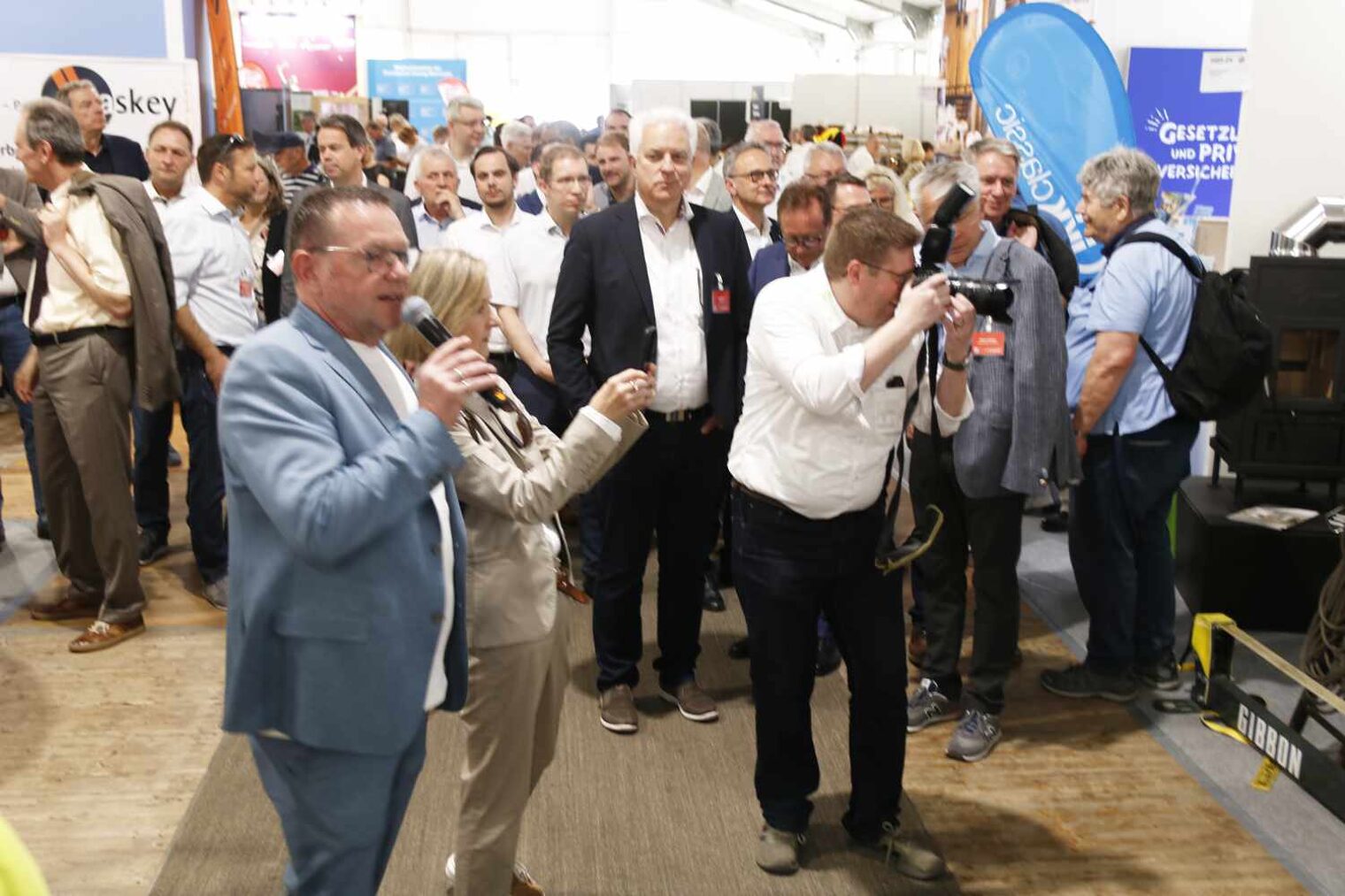 Zahlreiche Besucher beim Rundgang durch eine Messehalle, im Vordergrund ein Mann im hellblauen Anzug beim Sprechen ins Mikro, daneben einer der fotografiert