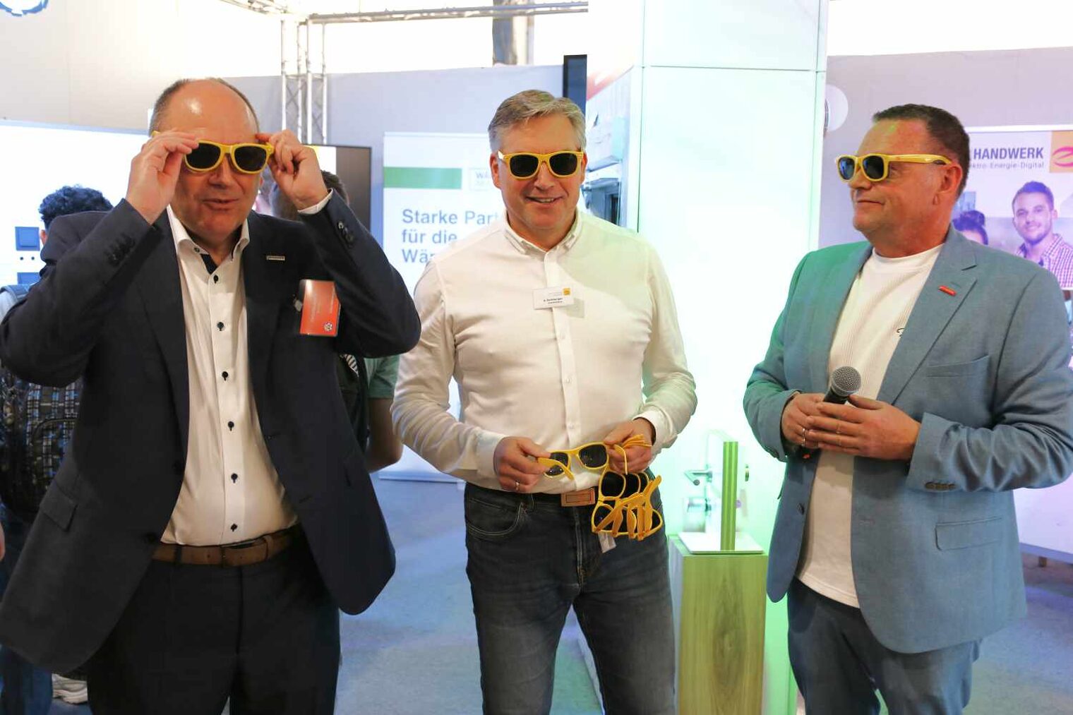 Drei Männer, der linke ist gerade im Begriff, sich eine gelbe Sonnenbrille aufzusetzen, die beiden anderen tragen bereits eine und lächeln