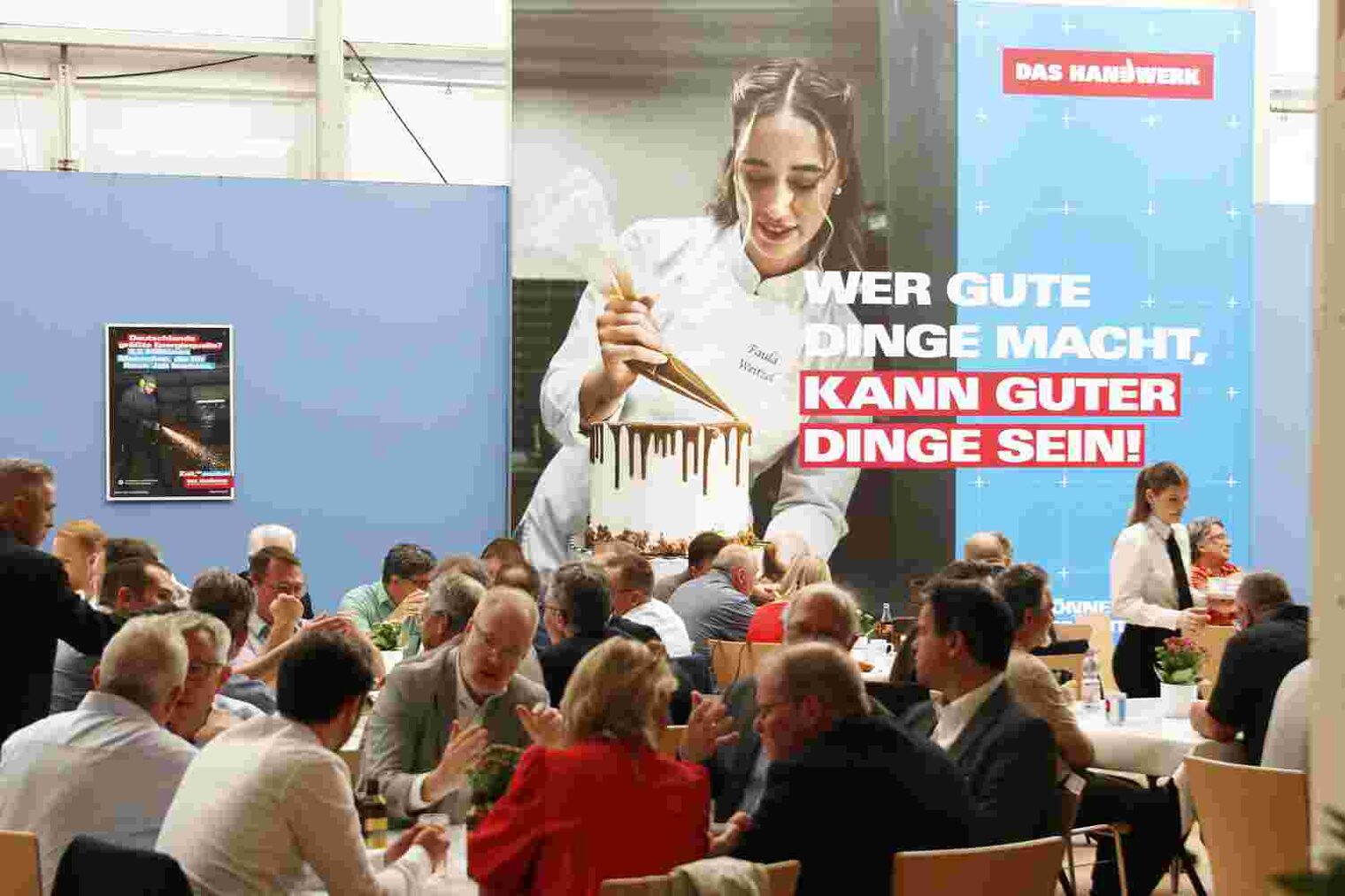 Personen, die in einer Messehalle im Bewirtungsbereich an Tischen sitzen und sich unterhalten, im Hintergrund ein großes Plakat mit einer Konditorin