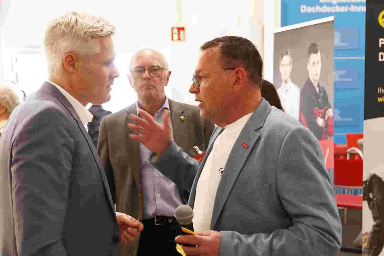 Drei Männer in einer Messehalle, der Mann rechts gestikuliert beim Gespräch mit dem Mann links, im Hintergrund beobachtet ein Mann das Geschehen