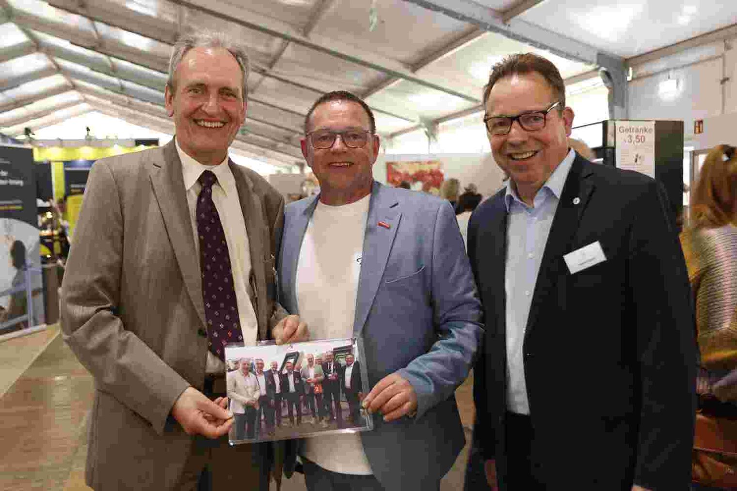 Drei ältere Männer in legeren Anzügen lachen in die Kamera, die beiden Männer links halten ein Bild, auf dem eine Gruppe von Personen zu sehen ist
