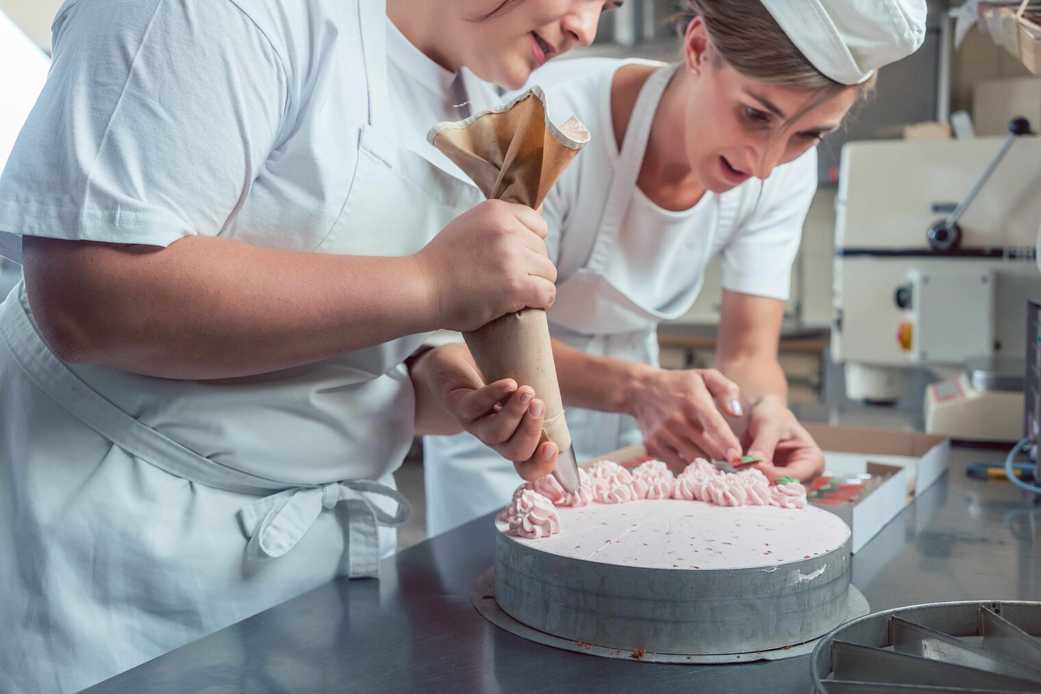 Zwei junge Konditorinnen dekorieren eine Torte, die linke Frau hat einen Spritzbeutel in der Hand und sprüht Cremetupfen auf, die rechte greift nach etwas