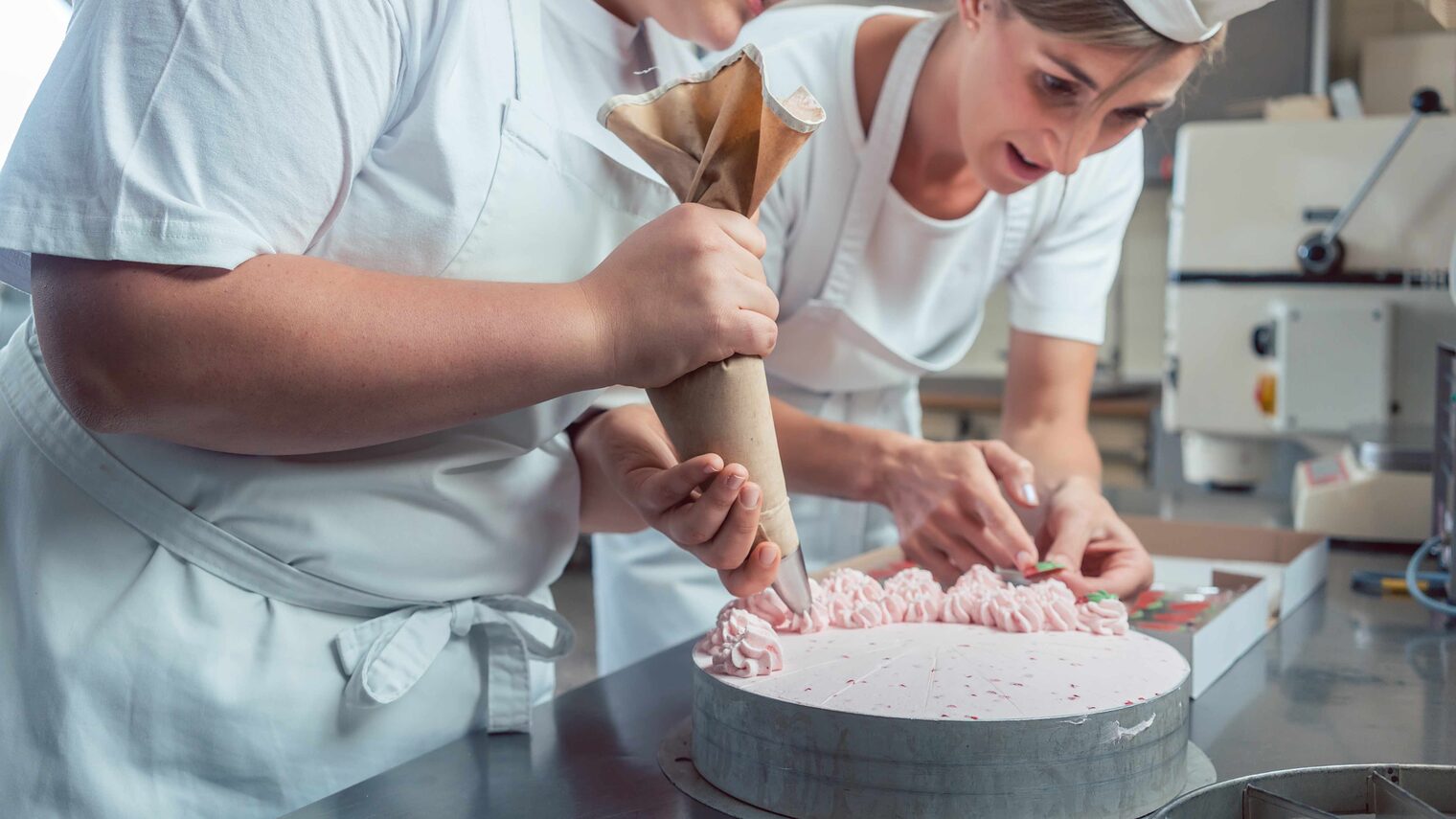 Zwei junge Konditorinnen dekorieren eine Torte, die linke Frau hat einen Spritzbeutel in der Hand und sprüht Cremetupfen auf, die rechte greift nach etwas
