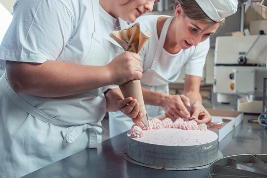Zwei junge Konditorinnen dekorieren eine Torte, die linke Frau hat einen Spritzbeutel in der Hand und sprüht Cremetupfen auf, die rechte greift nach etwas