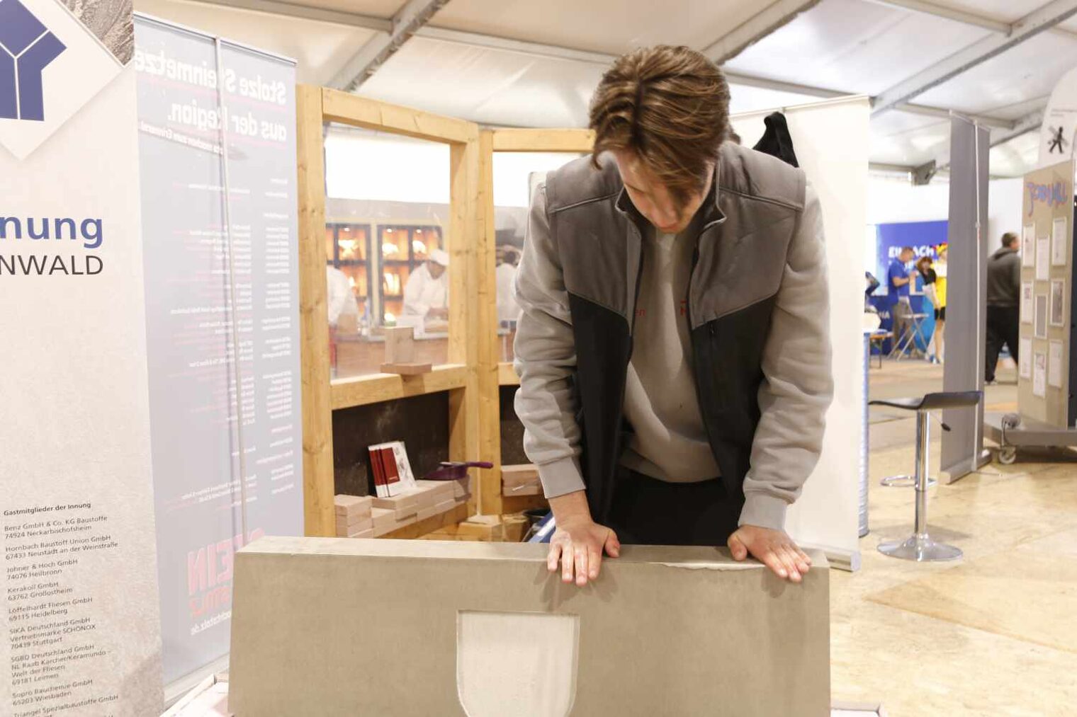 Junger Mann mit Weste auf einer Messe, der sich über eine geflieste Banklehne beugt und mit seinen Händen etwas festdrückt