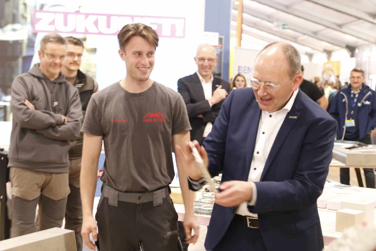 Mann hält Plakette in beiden Händen, auf die er begeistert schaut, im Hintergrund stehen andere Männer und lachen zufrieden