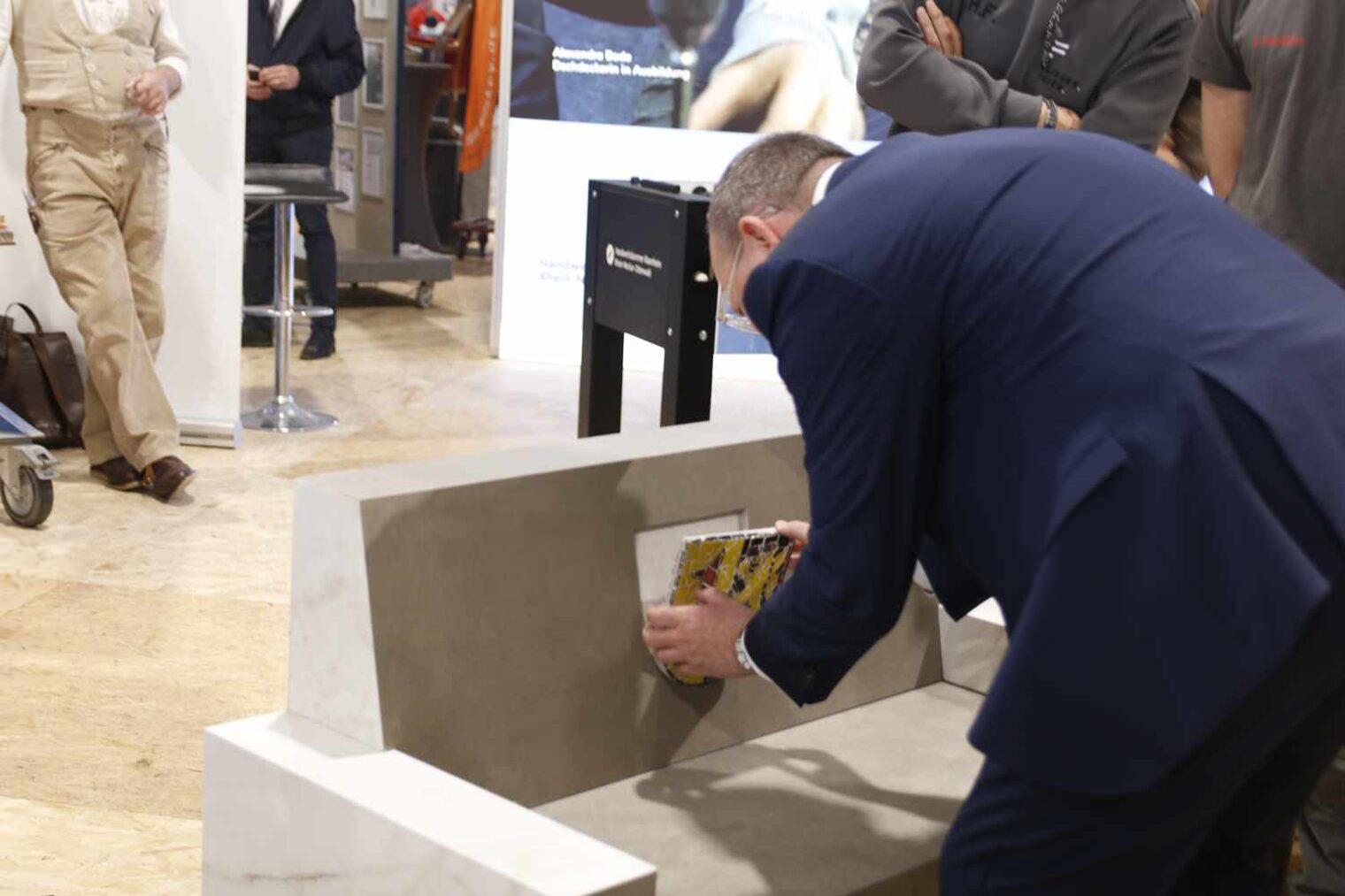 Mann mit dem Rücken zur Kamera beugt sich über eine Bank und setzt ein Wappen in eine Aussparung in der Lehne, im Hintergrund andere Menschen