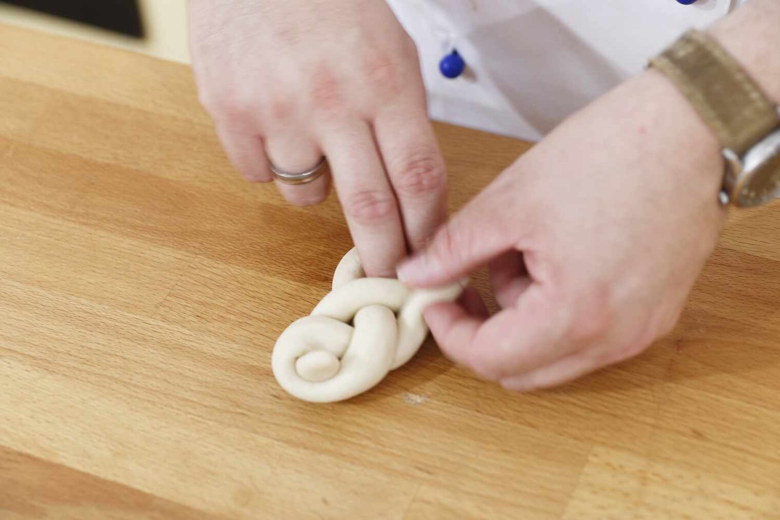 Ein Paar von Händen mit Ehering an der einen und Uhr an der anderen Hand formen aus Teig auf einer hölzernen Unterlage eine Zopfform
