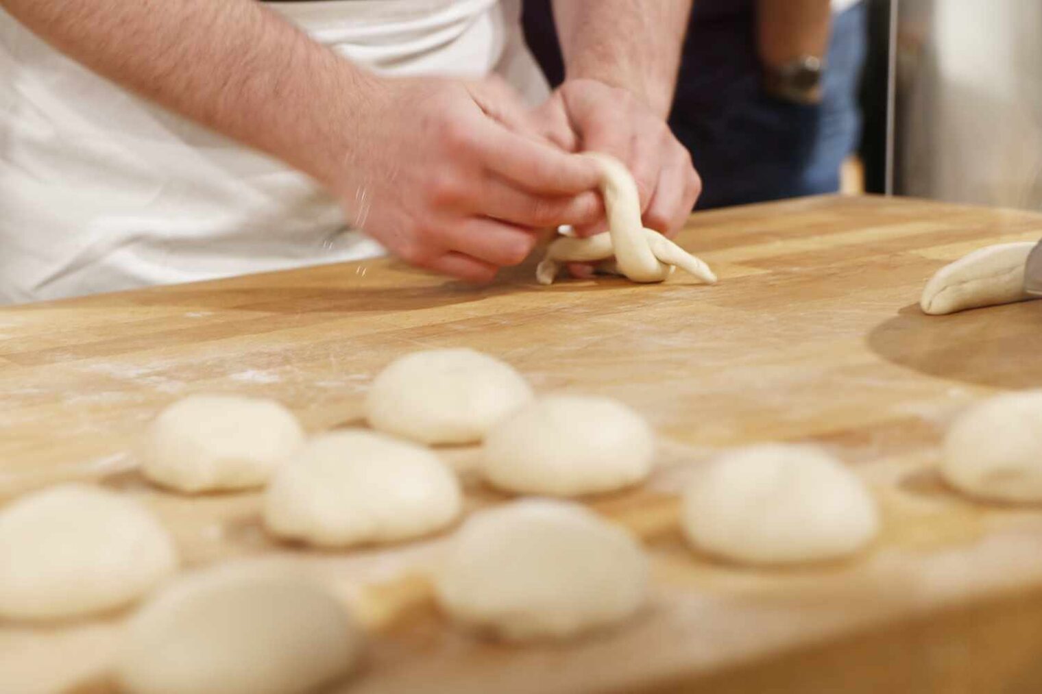Bemehlte Oberfläche aus Holz mit Teigkugeln in Brötchenform, hinten der Ausschnitt eines Bäckers im Bereich seiner Hände, die eine Zopfform flechten