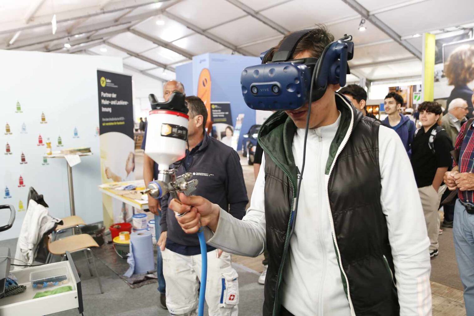 Jugendlicher trägt eine VR-Brille und führt in der rechten Hand eine Sprühpistole, im Hintergrund eine Messeszenerio mit weiteren Besuchern