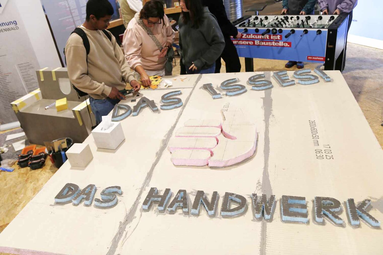 großflächige EXP-Bauplatte mit dreidimensionalem Schriftzug Das isses! Das Handwerk und Daumen, drei Personen hinten betrachten sie