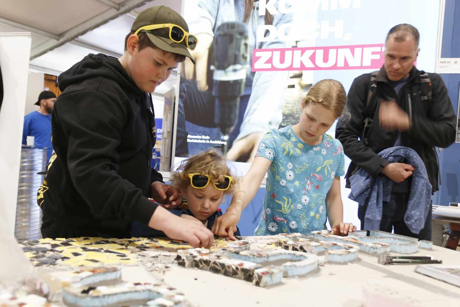 Drei Kinder in unterschiedlichem Alter und Größen stehen hinter einer Tafel und legen bunte Fliesenmosaik-Steine darauf, das Kinder in der Mitte kann gerade auf die Platte schauen