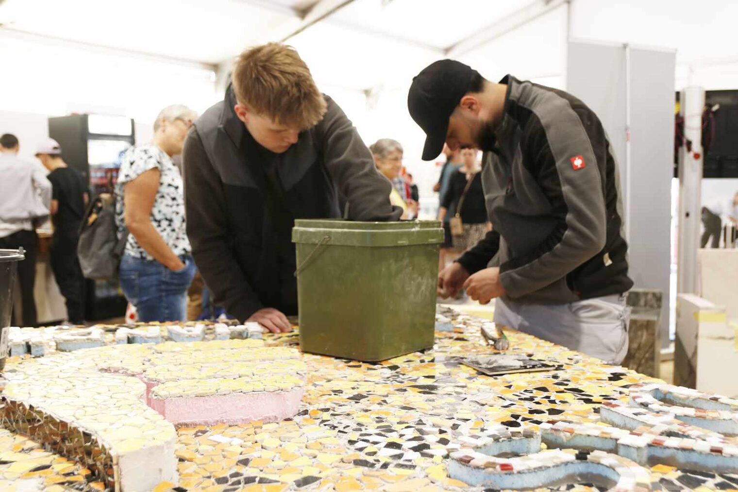 Im Vordergrund erstreckt sich ein mit bunten Mosaiken befliester Tisch ins Bild, hinten stehen zwei junge Männer und arbeiten daran, vor ihnen ein Eimer auf der Tafel