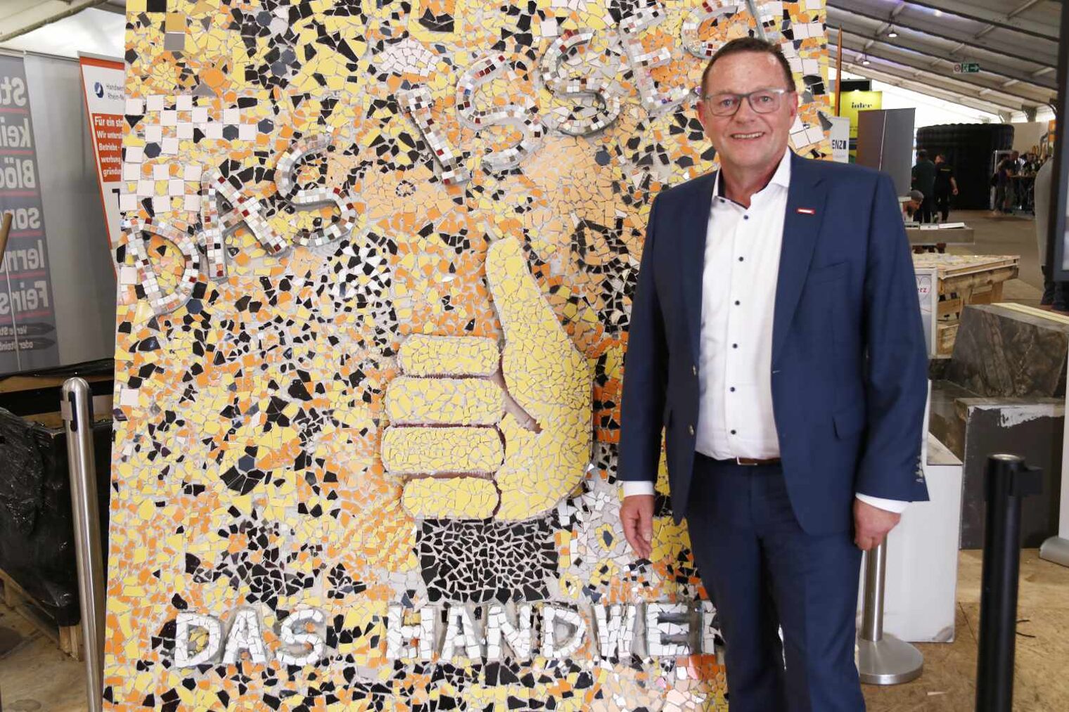 Mann im dunkelblauen Anzug mit Brille steht rechts eine mannhohen Tafel aus buntem Fliesenmosaik, auf dem mittig ein Daumsymbol ist und darunter Das Handwerk steht