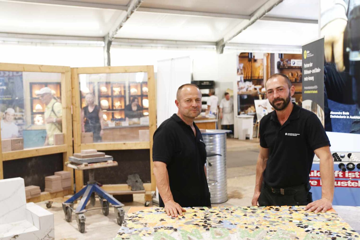 Zwei Fliesenleger stehen hinter einer aufgebockten Tafel, belegt mit bunten Fliesenmosaiken, beide haben ihre äußere Hand auf der Fläche liegen